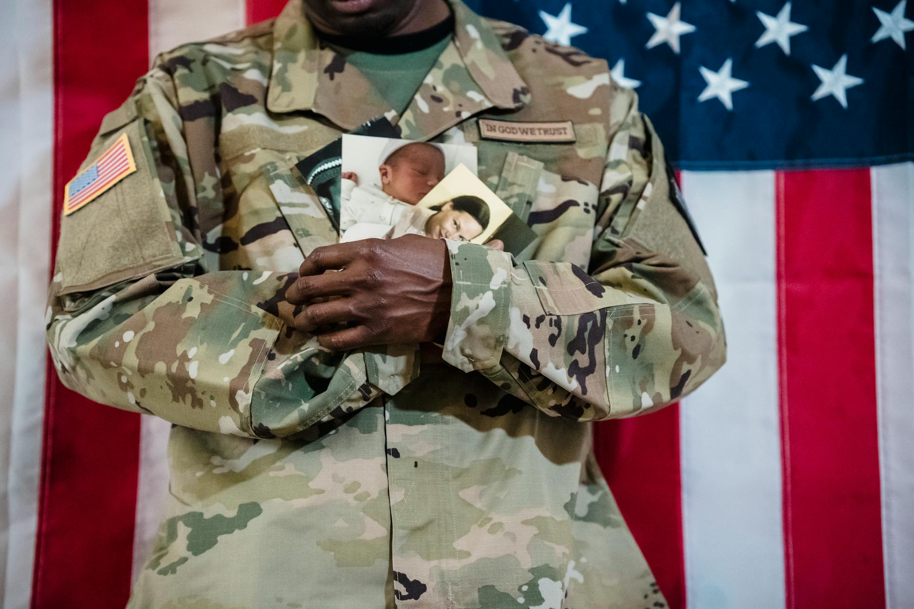 American soldier in uniform holding family photos in front of an American flag backdrop.