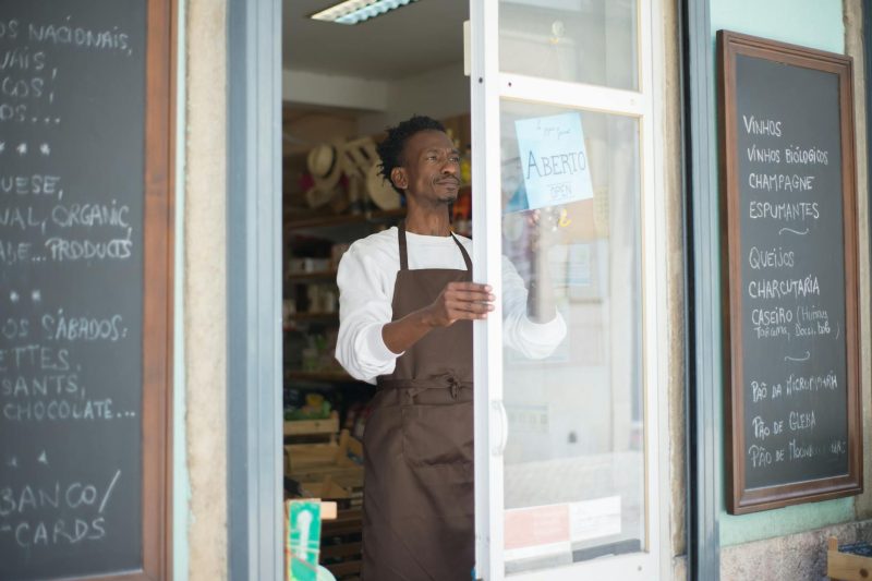 A man in an apron stands at the entrance of a grocery store, preparing to welcome customers.