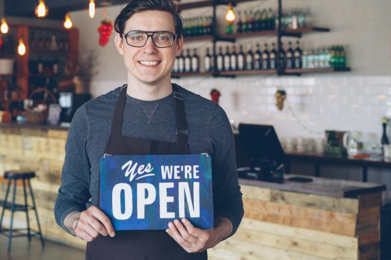 Barista smiles and welcomes customers in a cozy cafe with an open sign.
