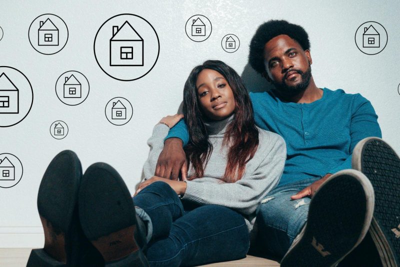 Couple sitting on the floor, dreaming about home ownership, surrounded by floating house icons.