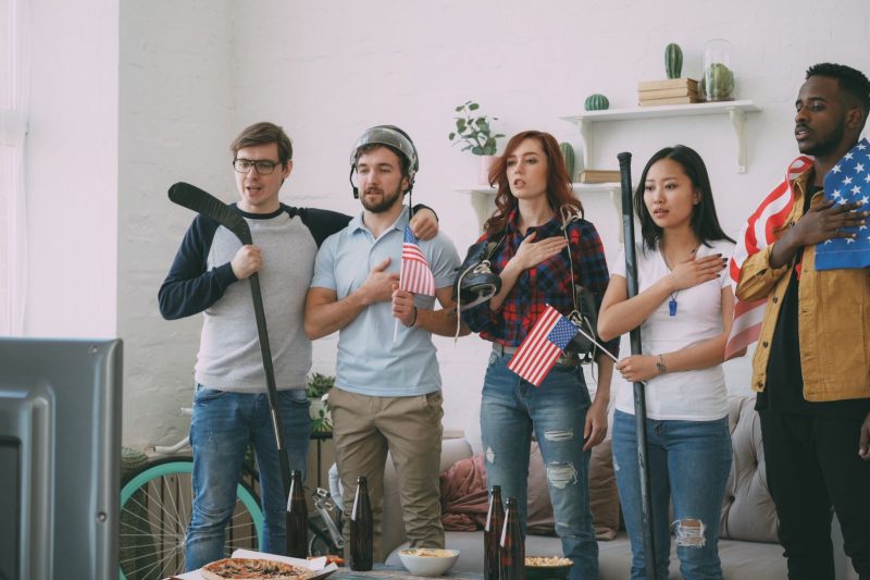 Diverse group of friends watching sports on TV, surrounded by American flags in a cozy living room.
