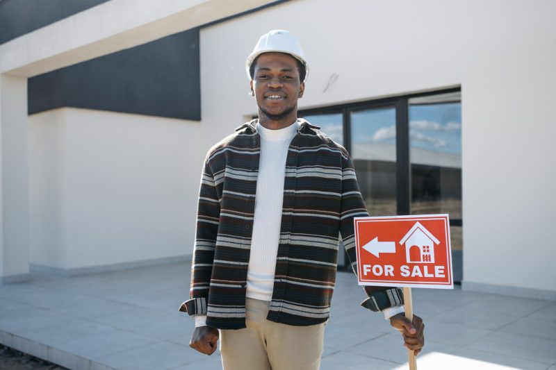 Young black male realtor smiling, holding a for sale sign in front of a modern house.