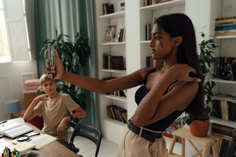 A woman takes a selfie in a stylish, plant-filled study with a young boy observing her.